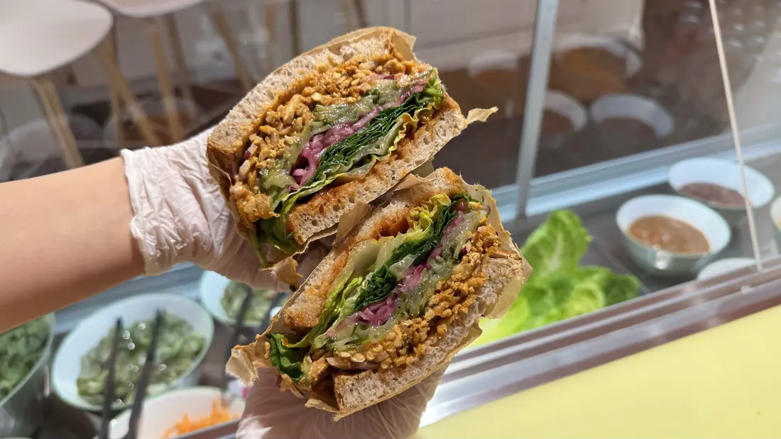 Sloppy Jo's plant-based deli sandwich on Meyers Bageri sourdough, cut in half and held up over the deli's prep counter — shredded tofu filling, pickled red onion, fresh lettuce and chili sauce visible, with fresh ingredients in the background